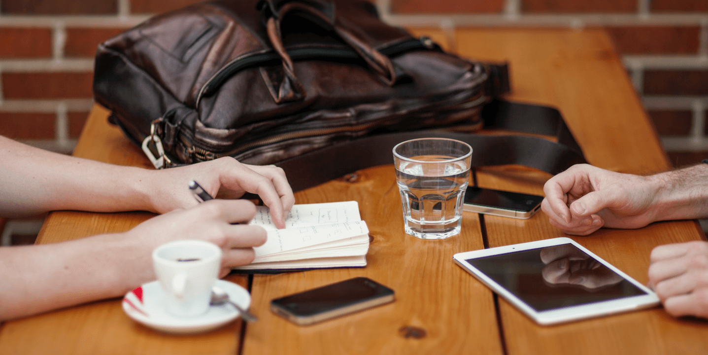 Two people at a table with a tablet, phones, a notepad and coffee