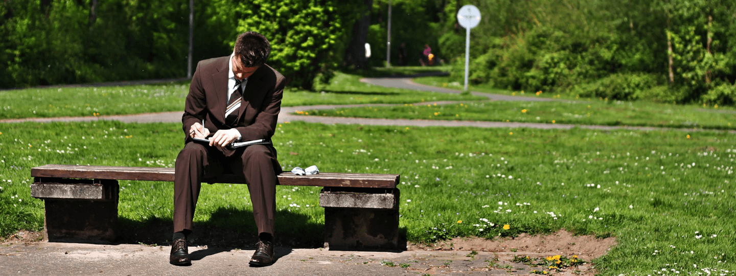 A man in a suite sitting on a park bench writing in a notebook