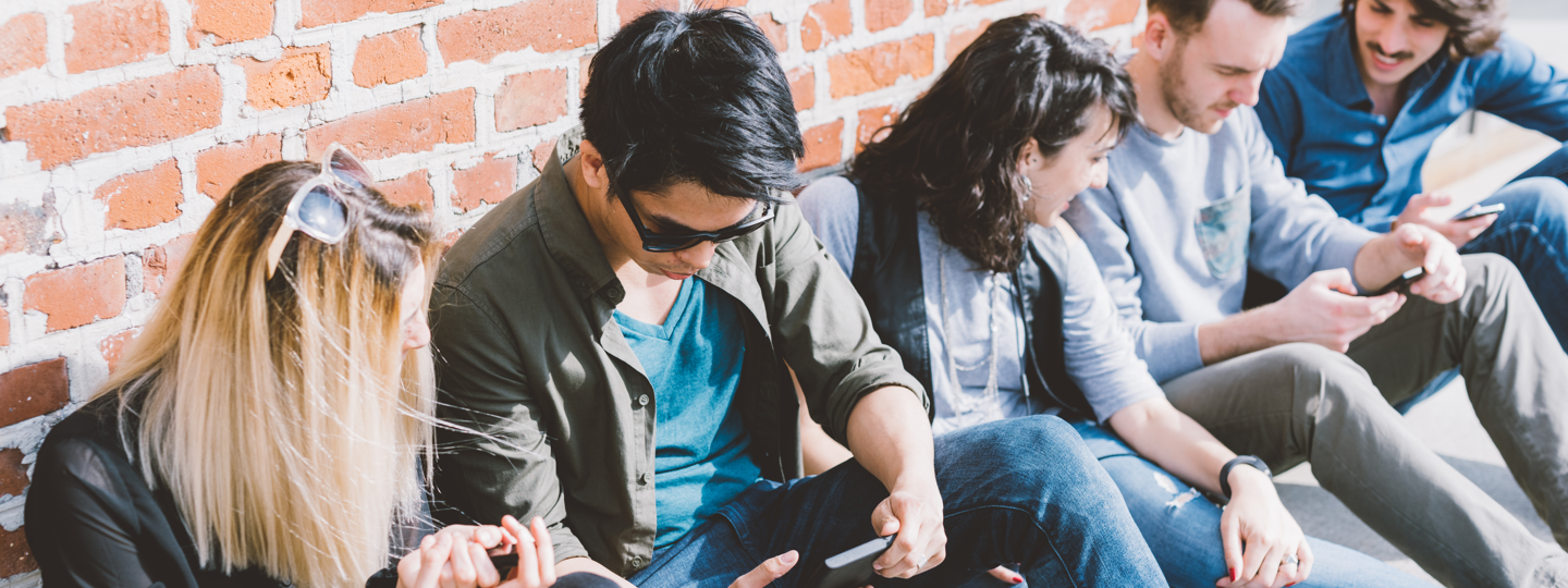 Five young people sitting against a brick wall looking at their phones