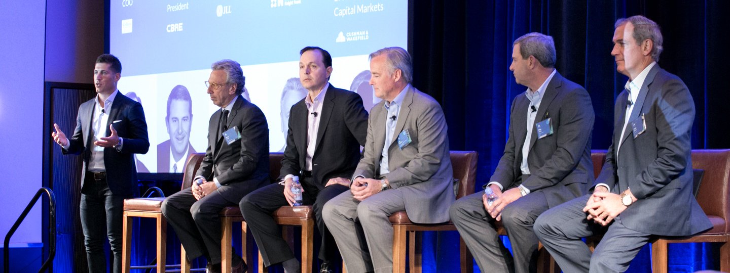 A man in a suit speaking next to 5 other panelists at a conference